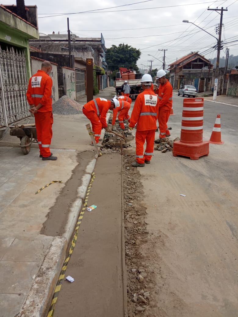 Seis funcionários trabalhando na reconstrução de sarjeta, isolados por cones na rua.